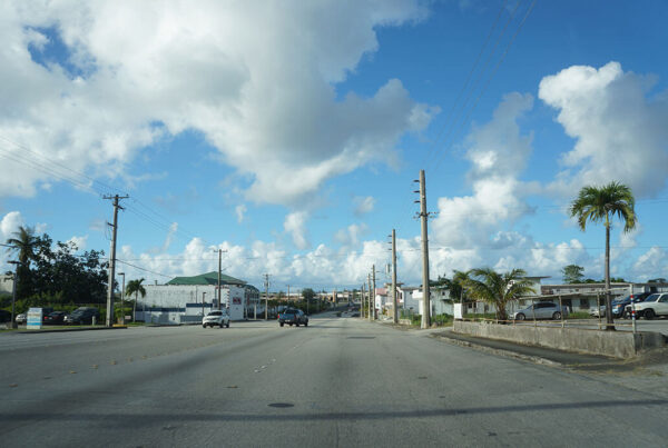 A street in Guam lined with power lines and palm trees under a cloudy sky.